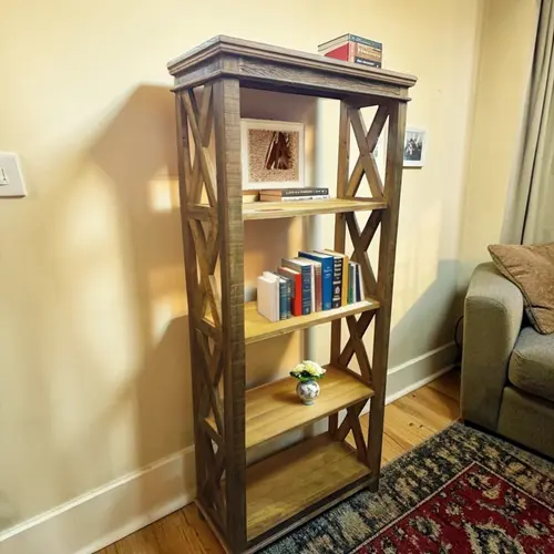 Estante livreiro alta de madeira de demolição com estilo rústico, design em X nas laterais e acabamento em tom natural, sem fundo e sem gavetas. Possui quatro prateleiras abertas ideais para organizar livros e objetos decorativos. Na imagem, a peça está decorada com livros, um vaso pequeno com flores e itens decorativos, posicionada em sala de estar ao lado de um sofá, sobre piso de madeira com tapete estampado. Perfeita para compor ambientes rústicos, industriais ou modernos com charme e personalidade.